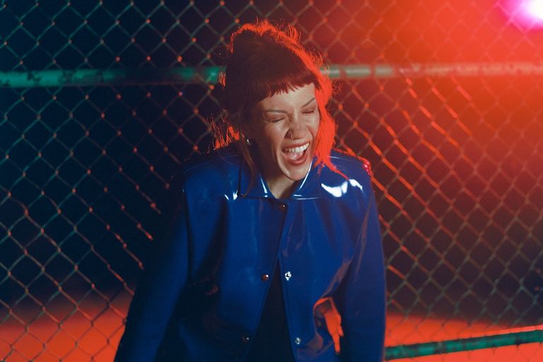 Singer-songwriter Bess Atwell smiling in a blue patent jacket against a chain-link fence under red lighting, photo by Elizabeth Landry.